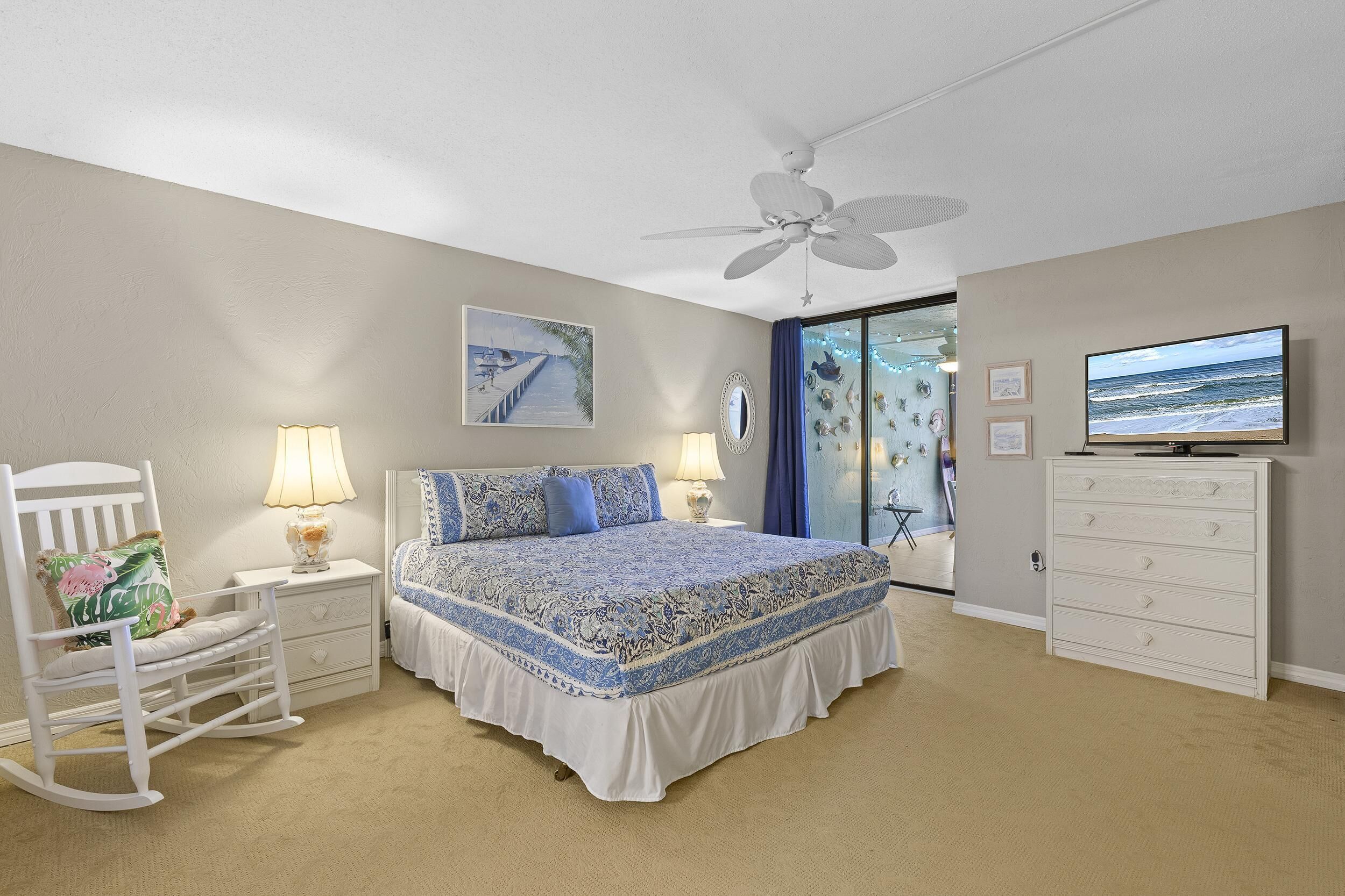 A bedroom with a bed, a rocking chair, a dresser, a ceiling fan, and a television.