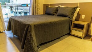 Un lit bien fait, recouvert d’une couverture sombre, une table de chevet avec un tiroir, et une vue sur les bâtiments par la fenêtre.