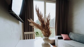Ein weißer Tisch mit einer Vase aus getrockneten Pflanzen, ein graues Sofa mit einem rosafarbenen Kissen und ein Fenster mit Vorhängen.