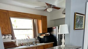 Eine Küche mit Holzschränken, einem schwarzen Kühlschrank, einer Mikrowelle und einem Spülbecken. Es gibt einen Deckenventilator, ein Fenster mit Vorhängen und einen Stuhl mit einem gewebten Griff.