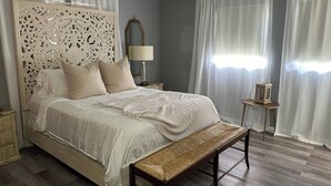 Ein Schlafzimmer mit einem großen Bett, einem Holz-Kopfteil, einem Nachttisch mit Lampe, einer Bank und einem kleinen Tisch mit einem dekorativen Gegenstand.