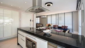 A modern kitchen with a large island, built-in oven, and a glass cooktop. - Cobertura em Jurerê a 200m da praia (Florianopolis)