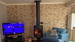 A living room with a stone-patterned wall, a black wood-burning stove, a blue sofa, and a television displaying a sports event.
