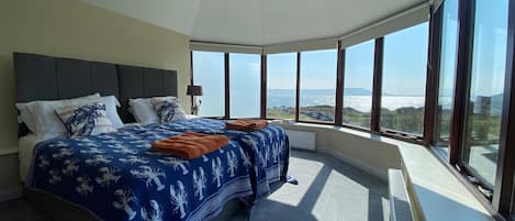 A bedroom with a curved ceiling, large windows, a bed with a blue floral bedspread, and a view of the sea.