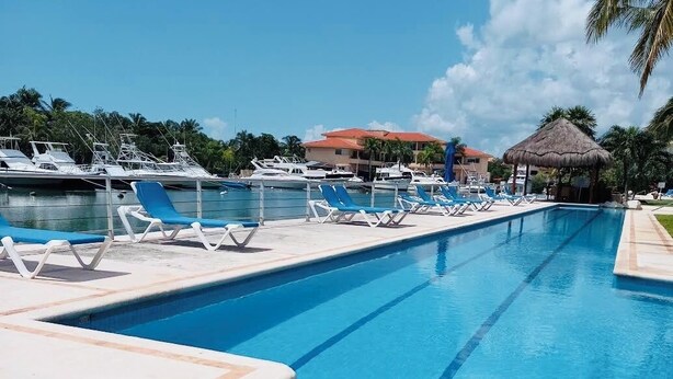 Ein Swimmingpool mit Liegestühlen, eine Reetdachhütte und Boote im Hintergrund.