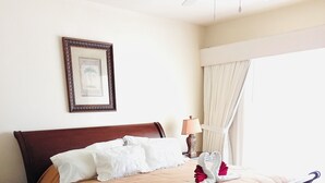 Ein ordentlich bezogenes Bett mit brauner Tagesdecke, weißen Kissen und einem Zierkissen mit roter Blüte. Darüber an der Wand hängt ein gerahmtes Bild, daneben steht eine Nachttischlampe und ein Fenster mit Vorhängen.