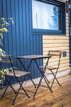A wooden deck with a table and chairs, a window, and a blue wall. - Tiny house with Nordic bath 10 minutes from the Baie de Somme (Saint-Blimont)