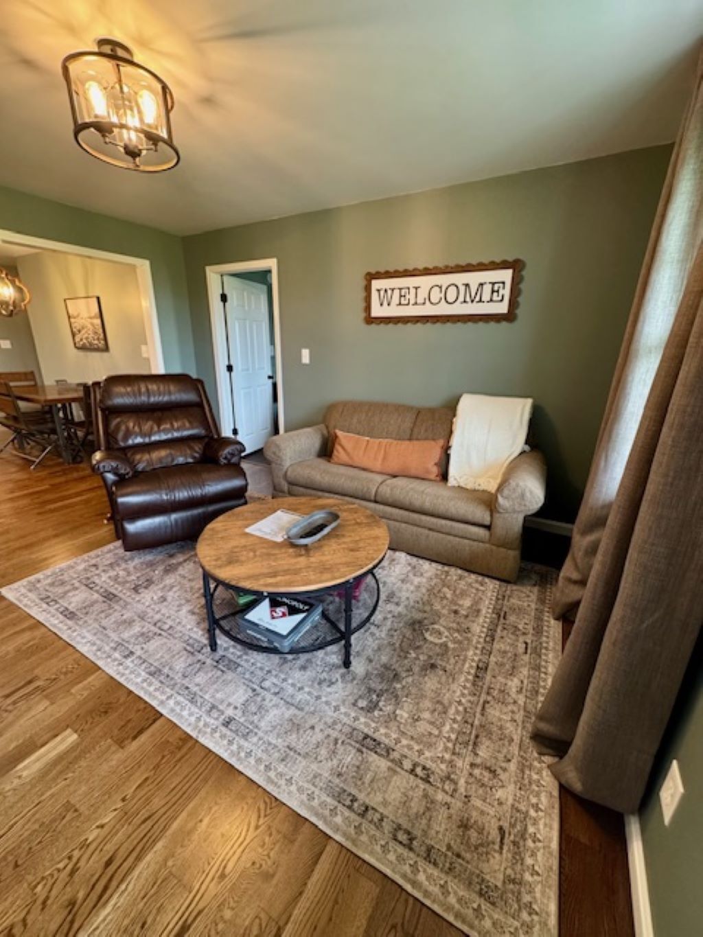 A living room with a leather chair, a sofa, a coffee table, and a 'WELCOME' sign on the wall.