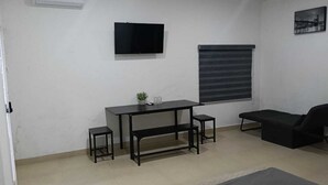 Una habitación con una mesa negra, dos taburetes negros, un banco negro, un sofá negro, un televisor montado y una ventana con persianas.