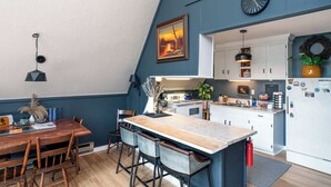Ett kök med ett matbord i trä, barstolar och en köksö. På väggen hänger en klocka och på kylskåpet sitter en krans.