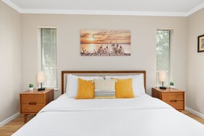 A bedroom with a bed, two bedside tables, a nightstand, a painting, and two windows with blinds.