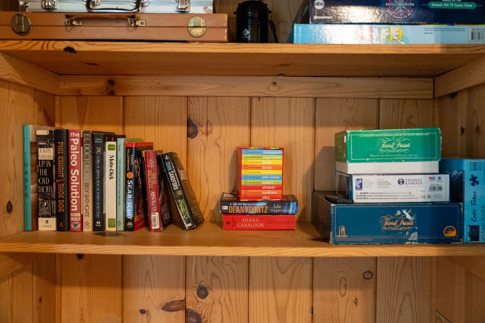 Una estantería de madera con varios libros, una pila de cajas de juegos y una maleta.