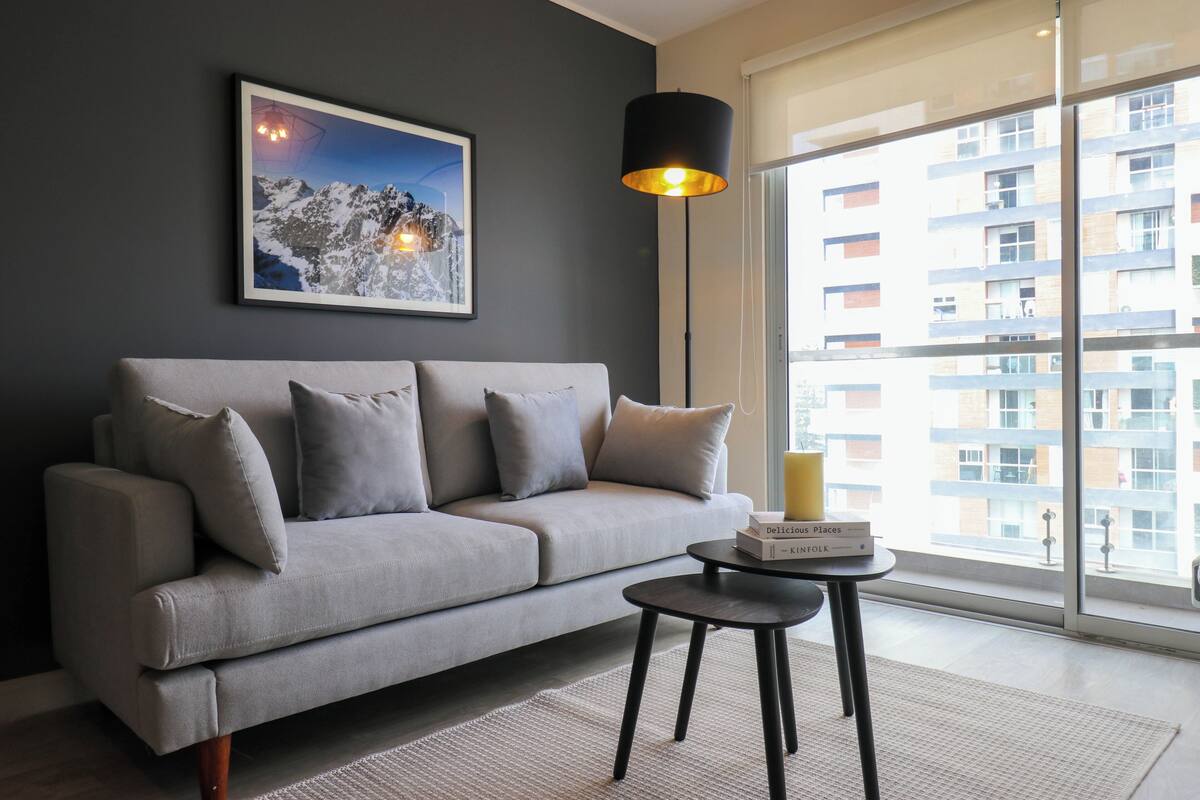 A modern living room with a grey sofa, a small round table, a floor lamp, and a large window with a view of a building.