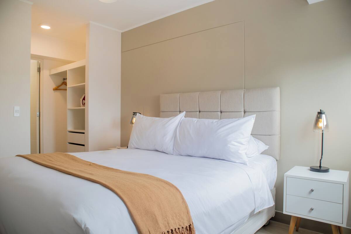 A neatly made bed with a white comforter and a brown throw blanket, a bedside table with a lamp, and a walk-in closet with shelves.