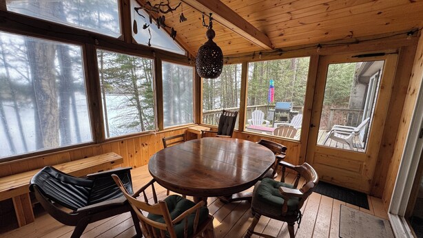 Interno di una baita in legno con un tavolo rotondo circondato da sedie, ampie finestre e vista sul bosco.