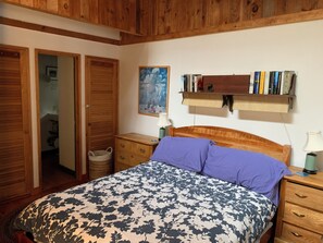 A bedroom with a wooden bed, a bedside table, a nightstand, a wall-mounted shelf with books, and a painting.