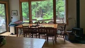 A room with a large wooden table and chairs, a fireplace, and a view of a forest through large windows.