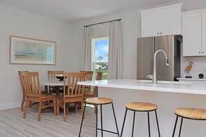 A modern kitchen with a stainless steel refrigerator, white cabinets, wooden chairs, and a large window with curtains.