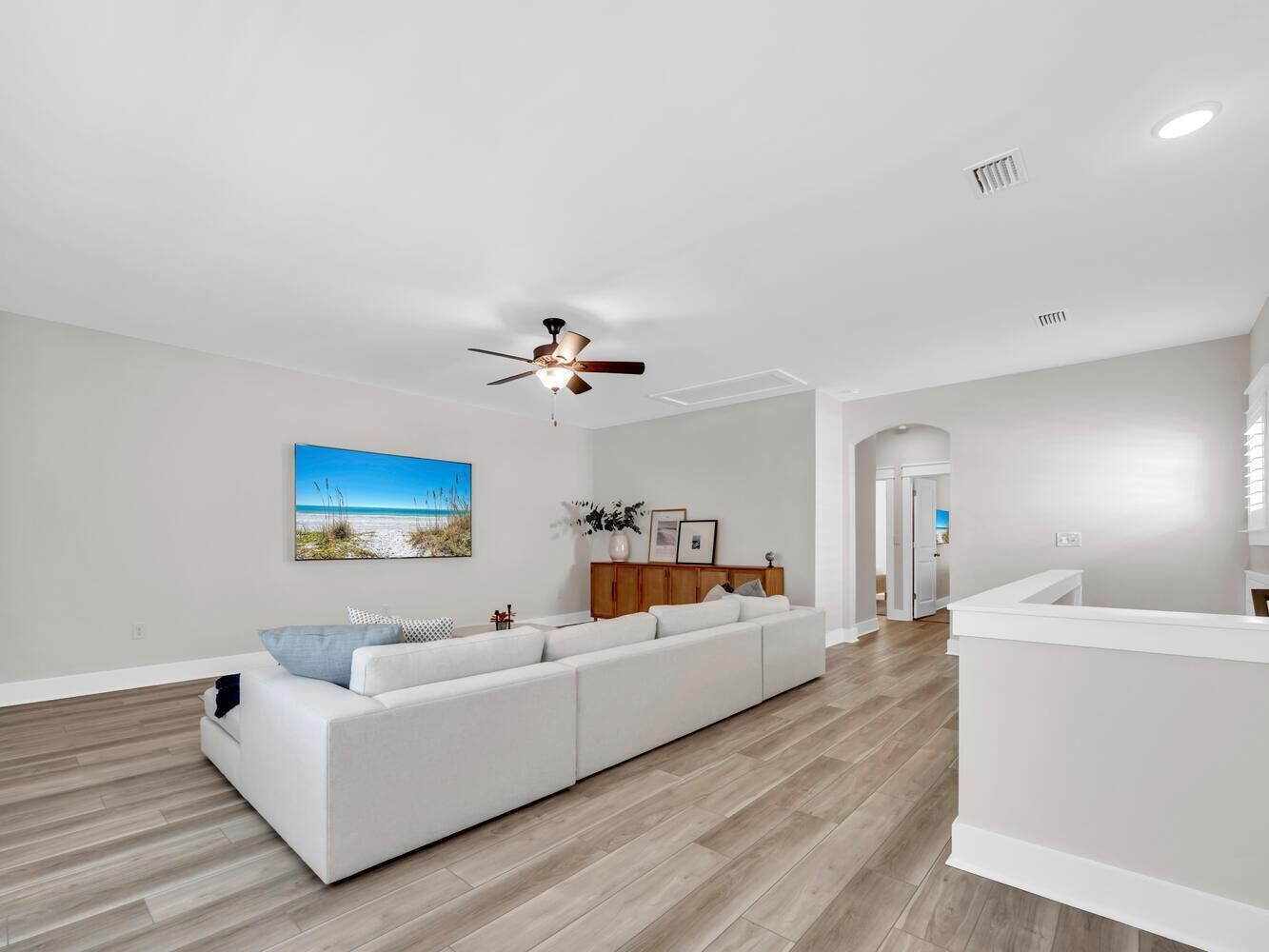Un salon moderne avec un canapé blanc, un plancher en bois, un ventilateur de plafond et un tableau représentant une scène de plage.