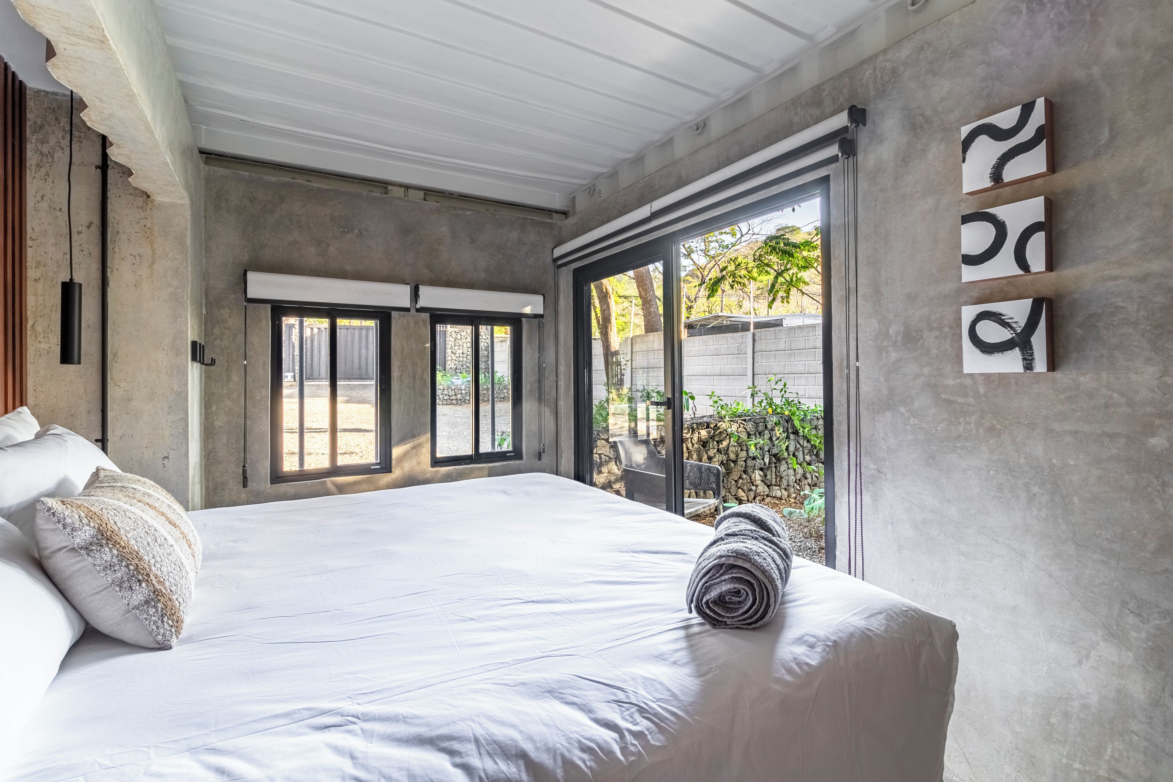 A bedroom with a large bed, a view of the outdoors through a sliding door, and a concrete wall with abstract art.