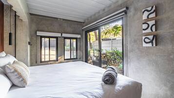 A bedroom with a large bed, a view of the outdoors through a sliding door, and a concrete wall with abstract art.