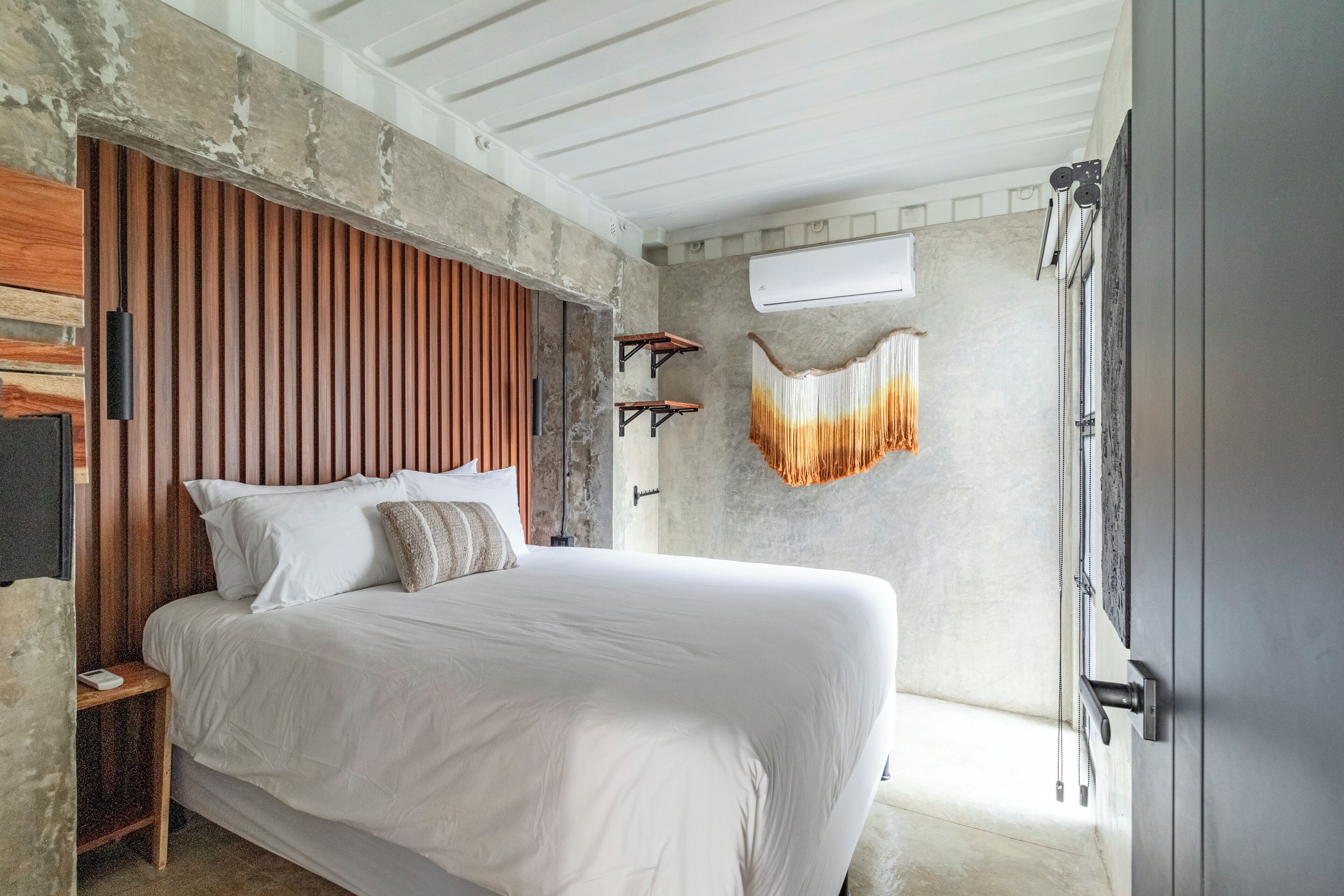 Ein Schlafzimmer mit einem großen Bett, einem Holzkopfende und einer Betonwand. Eine Pendelleuchte und ein kleiner Tisch befinden sich neben dem Bett.