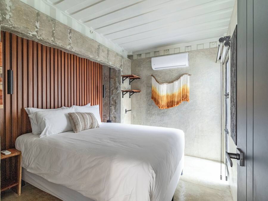 Ein Schlafzimmer mit einem großen Bett, einem Holzkopfende und einer Betonwand. Eine Pendelleuchte und ein kleiner Tisch befinden sich neben dem Bett.