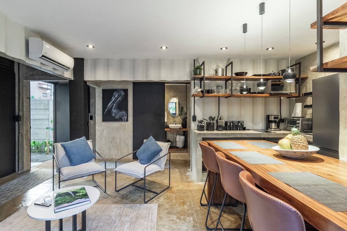Een moderne keuken met een houten eettafel, barkrukken en een keukeneiland. Er staat een stoel met kussen en een kleine tafel met een boek en afstandsbediening.