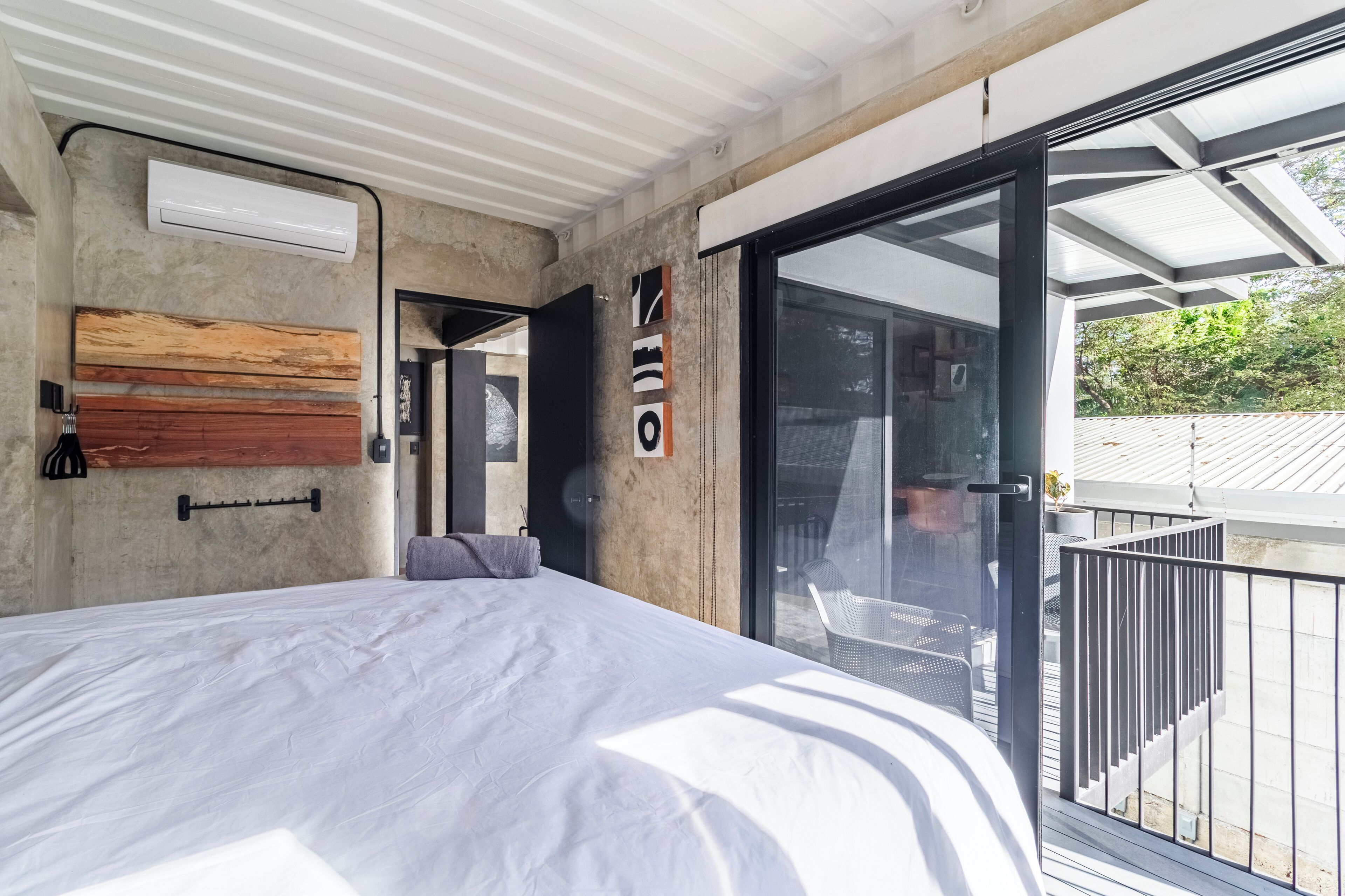 Une chambre moderne dotée d’un grand lit, d’une tête de lit en bois, d’un climatiseur mural et d’une porte-fenêtre coulissante donnant sur un espace extérieur avec un balcon.
