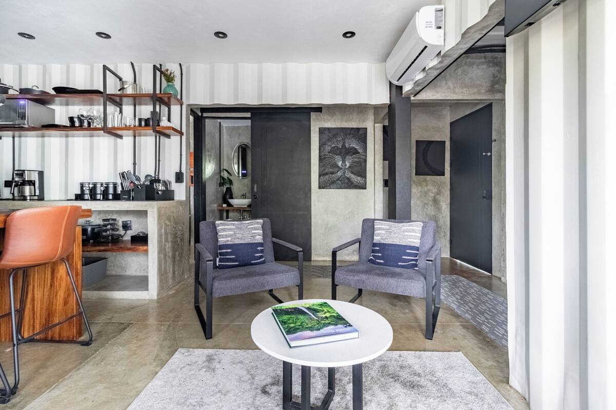 Een moderne keuken met een koffiezetapparaat, een eethoek met twee stoelen, een kleine tafel met een boek en een groot raam.