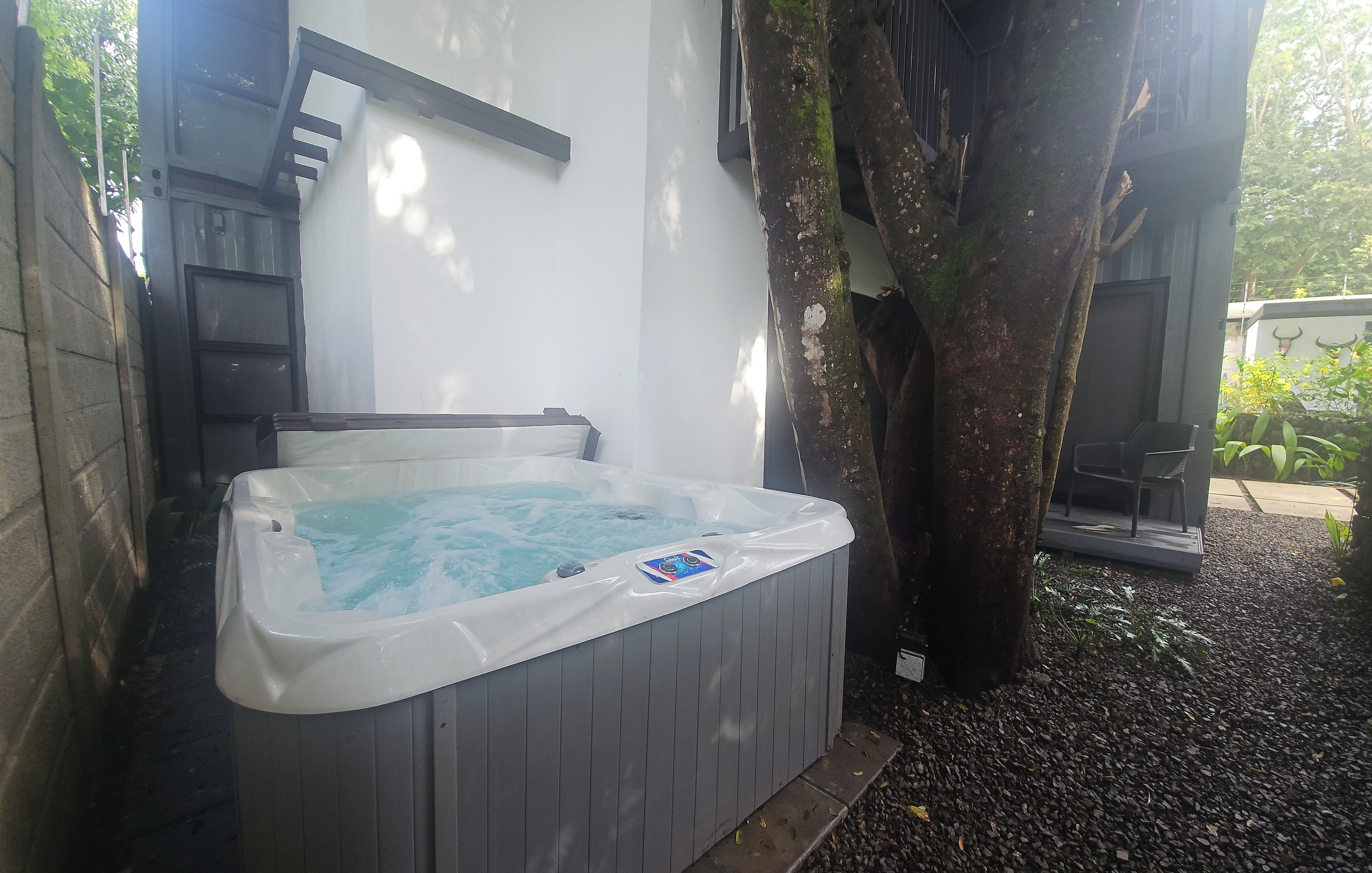 A hot tub next to a tree in a garden area with a white wall and a door in the background.