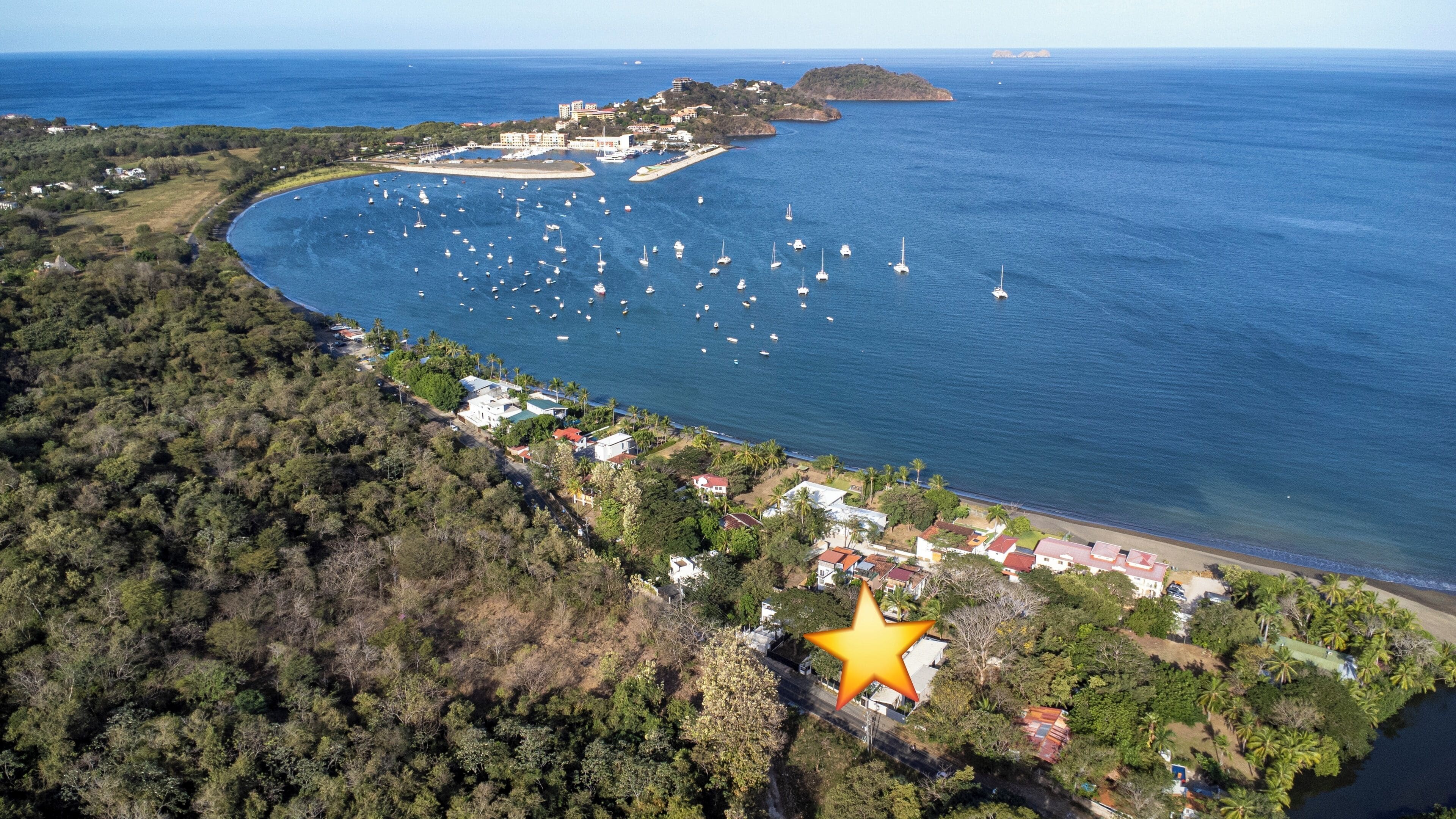 Une vue aérienne d’une zone côtière avec un port de plaisance, une végétation luxuriante et un bâtiment jaune imposant.