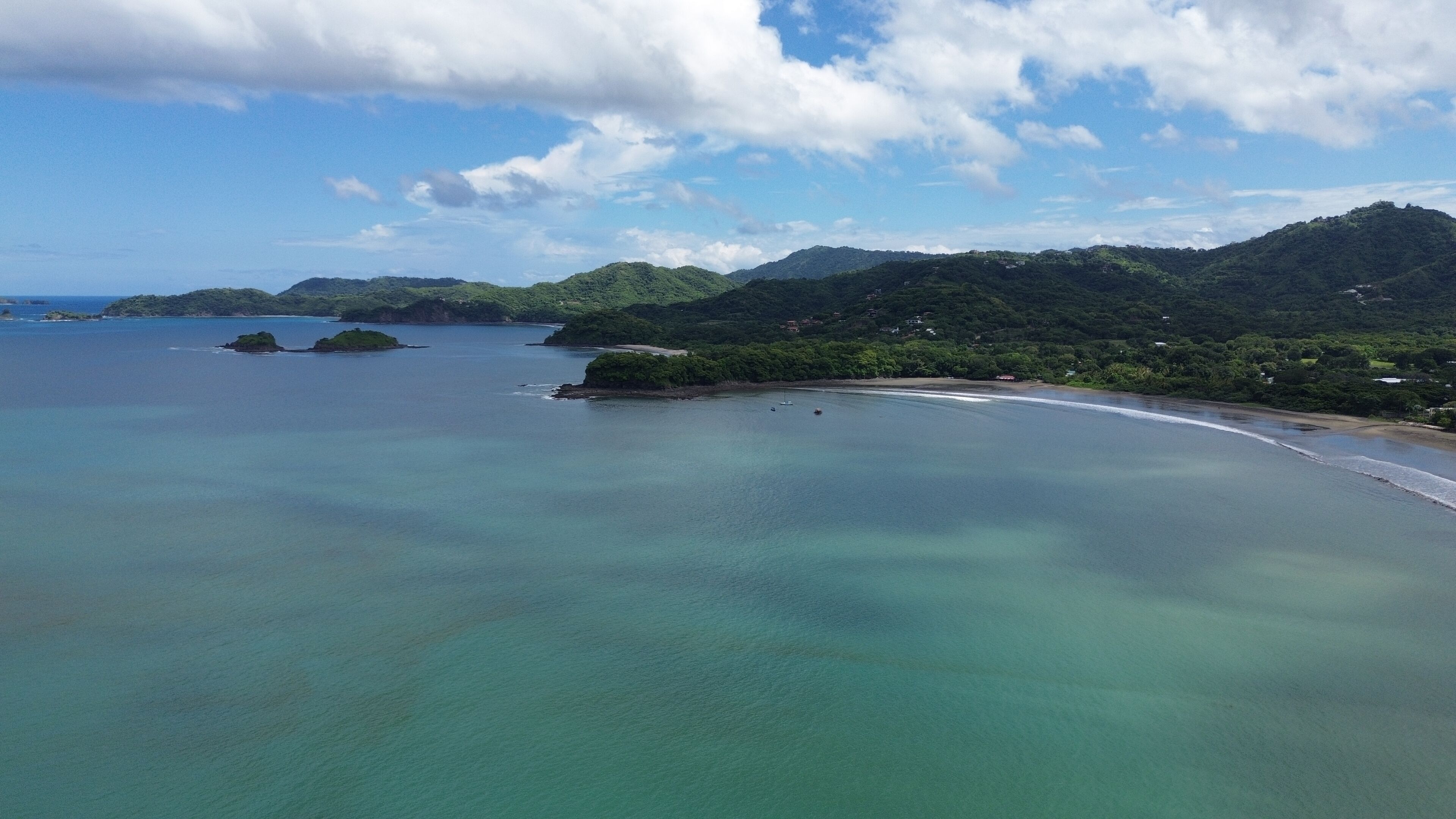 Paesaggio costiero con acque limpide, una spiaggia sabbiosa e dolci colline verdeggianti.