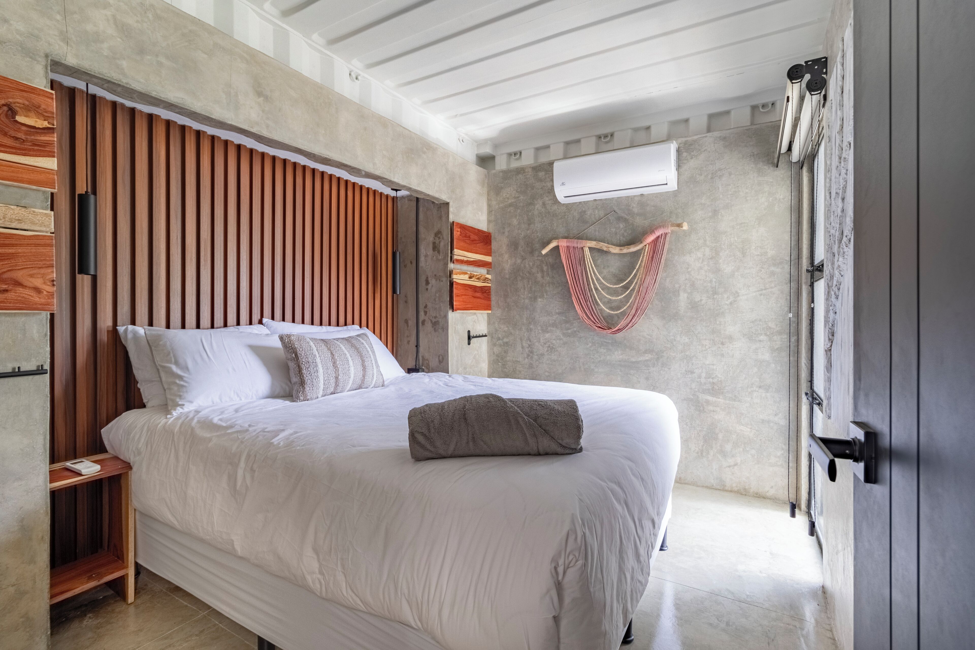 Une chambre moderne avec une tête de lit en lattes de bois, un grand lit avec une literie blanche, une table de chevet et un climatiseur fixé au mur.