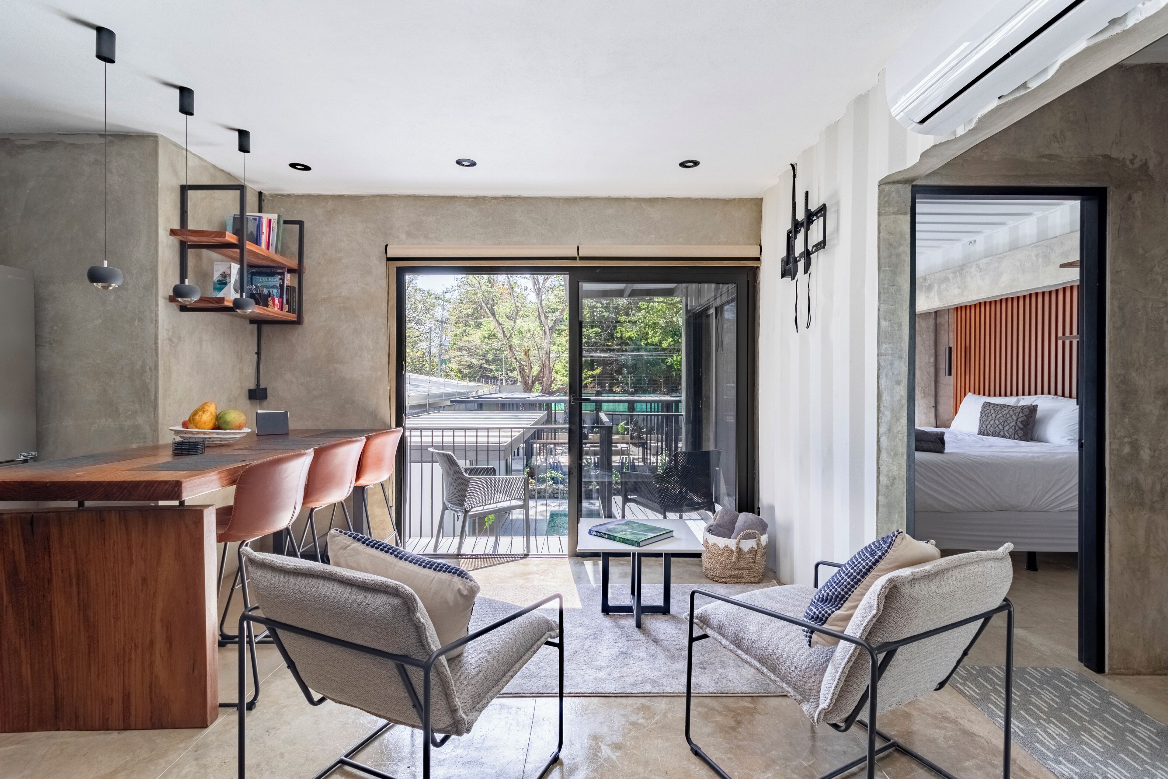 A modern interior with a wooden bar, two upholstered chairs, a small table, and a balcony with a view of the city.