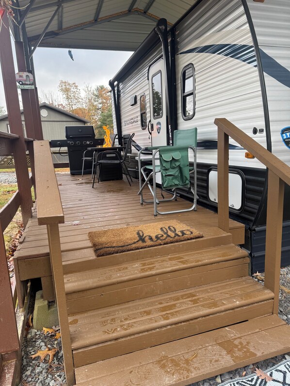 A covered deck area with a wooden staircase leading to a camper. - Cozy 2-bedroom RV in amazing Pipestem with AC, WiFi and Heat Smart TV (Pipestem)
