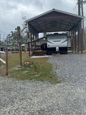 A covered RV parking area with a 'Welcome' sign, a 'Happy Camper' sign, and a 'Springdale' RV. - Cozy 2-bedroom RV in amazing Pipestem with AC, WiFi and Heat Smart TV (Pipestem)
