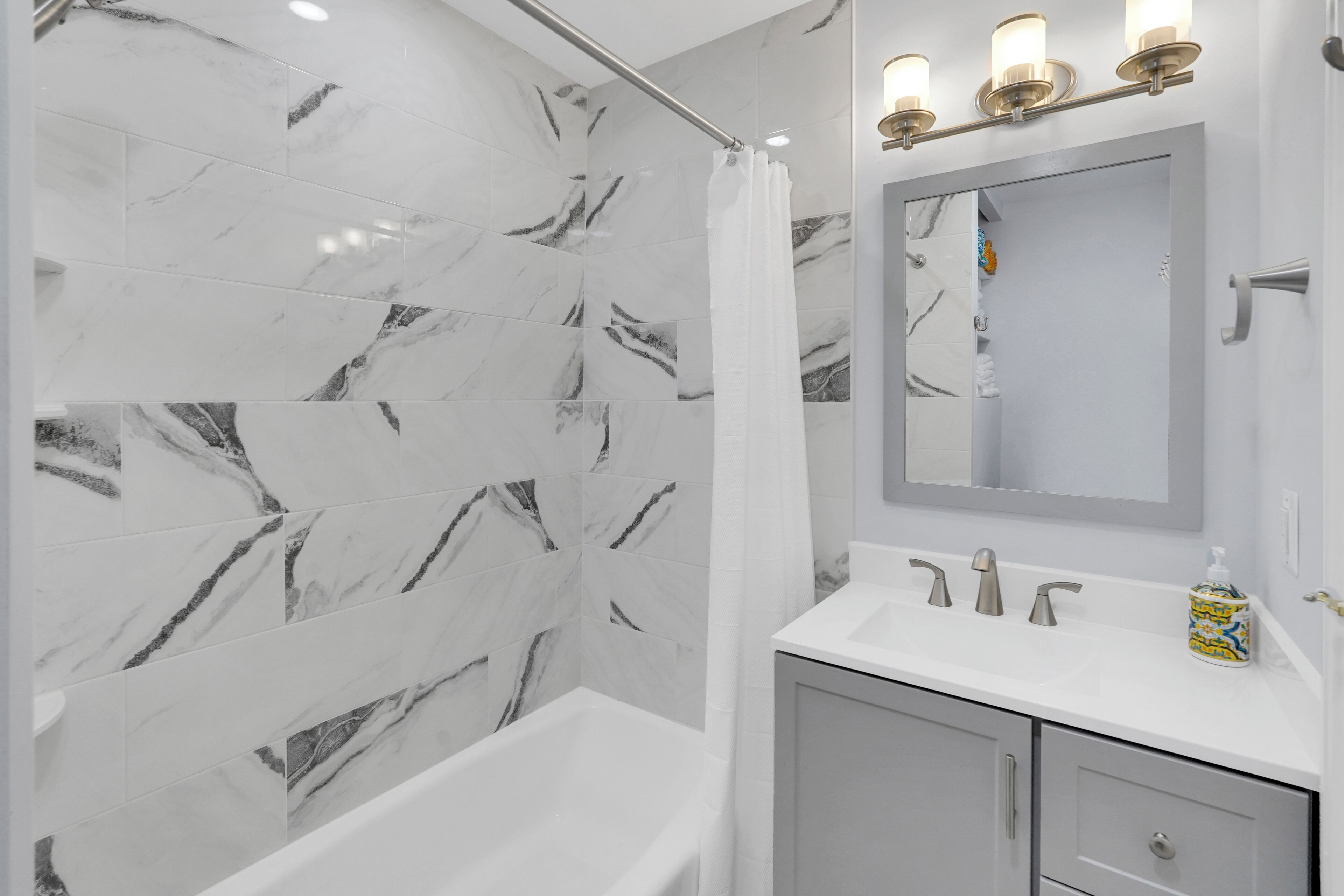 A modern bathroom with marble tiles, a bathtub, a vanity with a mirror and lights, and a shower curtain.