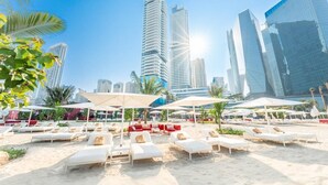A beach with sun loungers and umbrellas, palm trees, and modern high-rise buildings in the background. - Five Luxe JBR (Dubai)