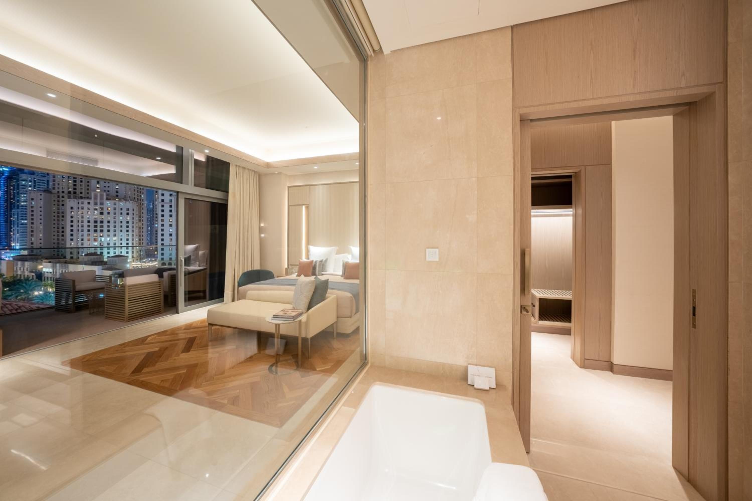 A modern hotel room with a large window, a sitting area with a sofa and a coffee table, and a separate bedroom with a bed and a walk-in closet.