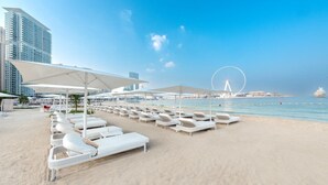 Ein Strand mit weißen Liegestühlen und Sonnenschirmen, einem Riesenrad und einem Kreuzfahrtschiff im Hintergrund.