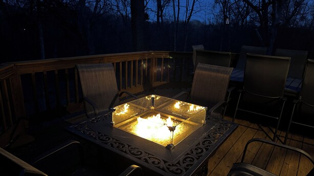 A deck with a fire pit in the center, surrounded by chairs and a wooden railing.