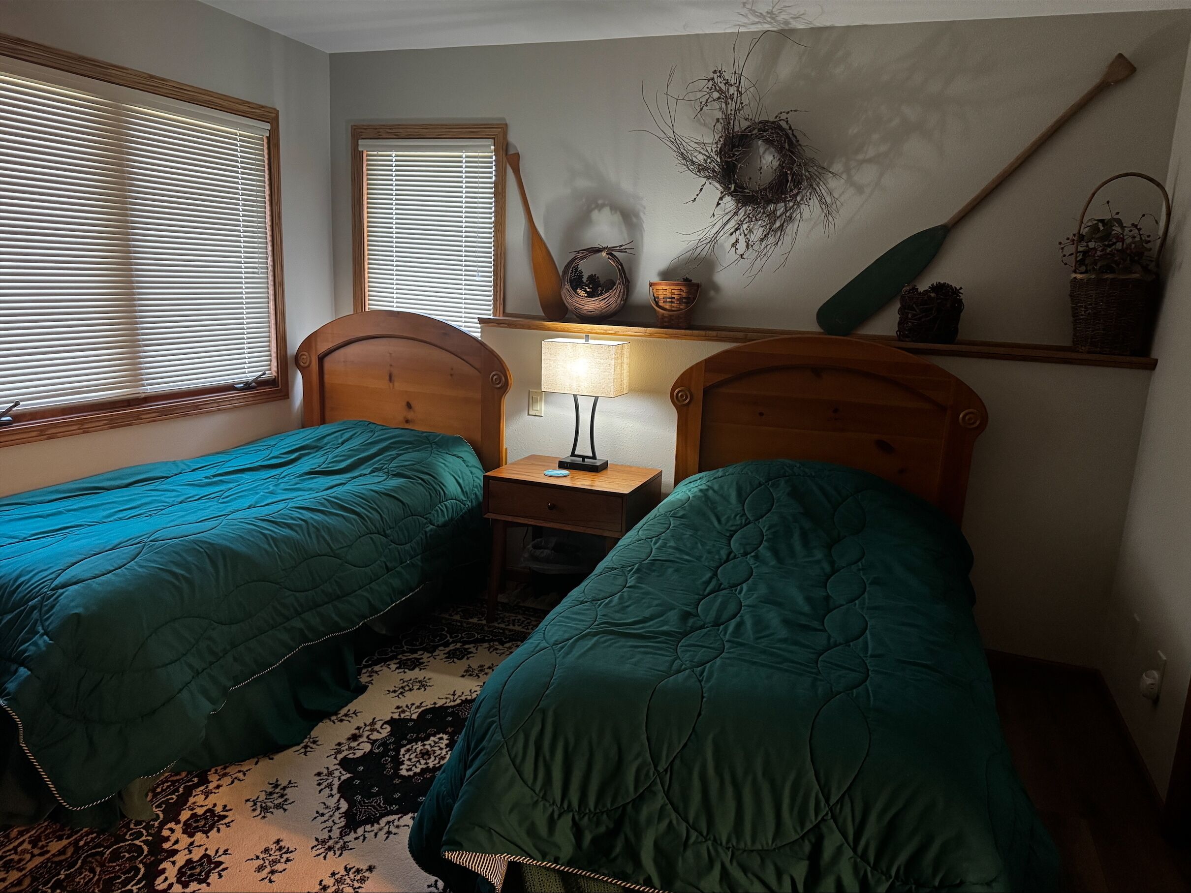 A bedroom with two single beds, a nightstand with a lamp, a shelf with decorative items, and a window with blinds.
