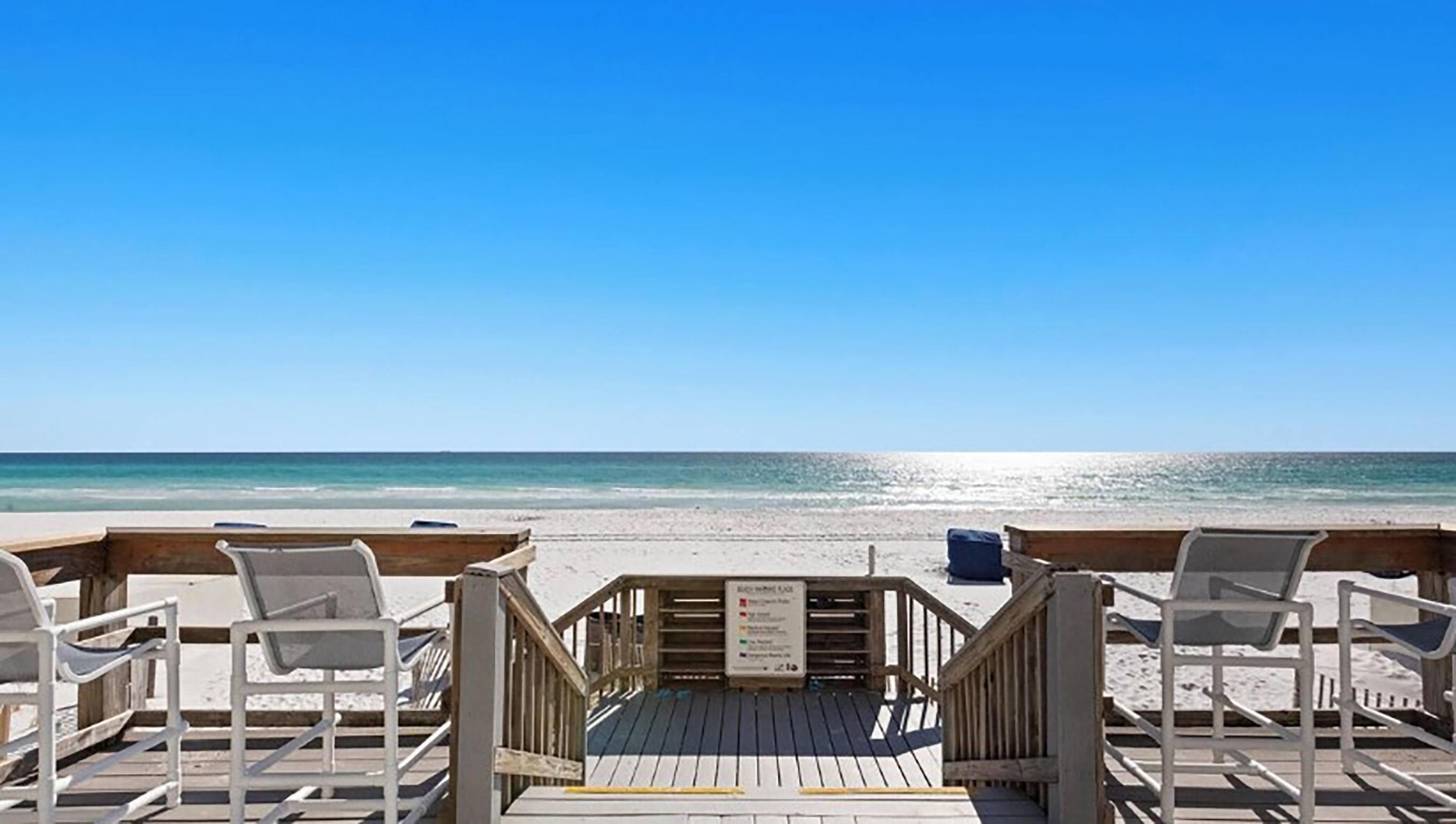 Une terrasse en bois, dotée de chaises de plage et d’un panneau indicateur, offre une vue sur une plage aux eaux d’un bleu limpide.