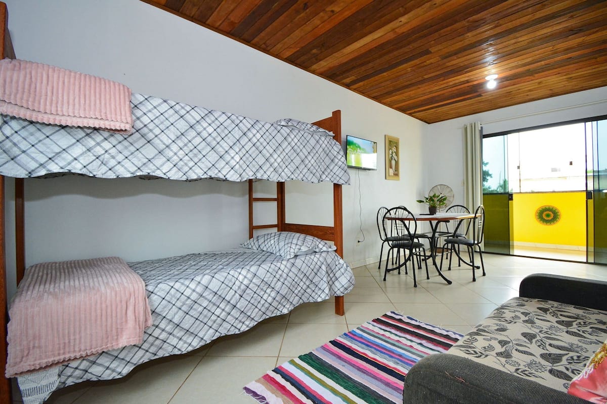Ein Zimmer mit einem Etagenbett, einem Esstisch mit Stühlen, einem kleinen Tisch mit einer Pflanze und einem bunten Teppich.