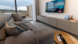 Moderni olohuone, jossa on suuri ikkuna, kuviollisilla koristetyynyillä varustettu sohva, seinälle asennettu televisio ja kaukosäädin puisen sivupöydän päällä.