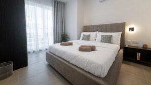 Moderni hotellihuone, jossa on suuri sänky, yöpöytä, seinälle asennettu ilmastointilaite ja näkymä ulos ohuiden verhojen läpi.