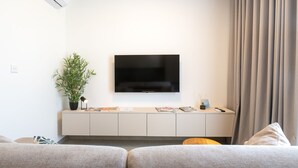 Ett modernt hotellrum med en platt-TV som är monterad på väggen, ett litet skåp med tidskrifter, en krukväxt och ett stort fönster med gardiner.
