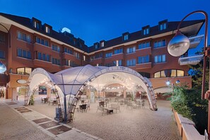 Un espacio de estar al aire libre con techo, con mesas y sillas, rodeado por un edificio de varios pisos con balcones.