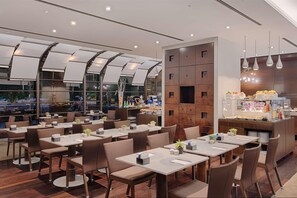Un restaurante moderno con varias mesas y sillas, una vitrina de vidrio con productos alimenticios y un ventanal que ofrece vistas de la ciudad.